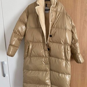 Puede incluir: Una larga chaqueta acolchada beige con un acabado brillante. La chaqueta tiene un forro de piel sintética y un cierre de botón.