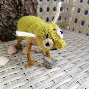 May include: A green knitted toy with large eyes and white antennae. The toy is standing on a white wicker surface.