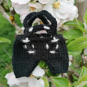 Könnte beinhalten: Eine kleine schwarze Stricktasche mit weißen Nähten, die an einem Zweig eines blühenden Apfelbaums hängt.
