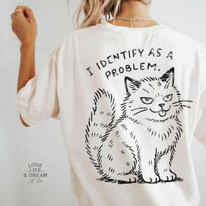 Puede incluir: Camiseta color crema con un gr&aacute;fico negro de un gato y el texto "I IDENTIFY AS A PROBLEM." La ilustraci&oacute;n del gato tiene una expresi&oacute;n traviesa. La camiseta tambi&eacute;n tiene el texto "LOVE LIFE & DREAM Art Shop" en la parte inferior.