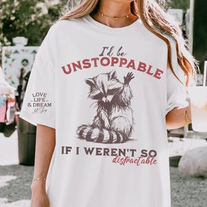 Może przedstawiać: Biała koszulka z ilustracją szopa pracza i napisem "I'd be UNSTOPPABLE IF I WEREN'T SO distractable." Projekt wykorzystuje brązowe i czerwone odcienie. Koszulka ma r&oacute;wnież napis "LOVE LIFE & DREAM Art Shop" po lewej stronie.