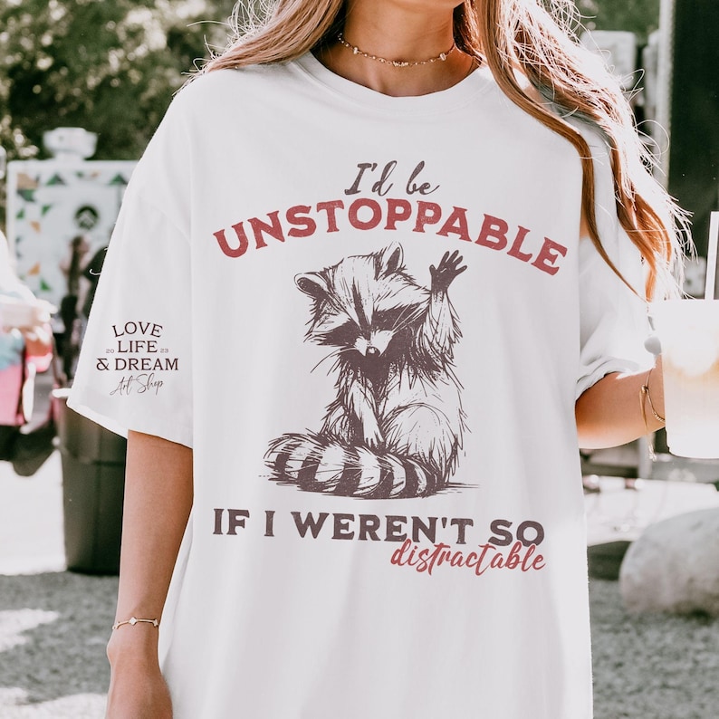 Może przedstawiać: Biała koszulka z ilustracją szopa pracza i napisem "I'd be UNSTOPPABLE IF I WEREN'T SO distractable." Projekt wykorzystuje brązowe i czerwone odcienie. Koszulka ma r&oacute;wnież napis "LOVE LIFE & DREAM Art Shop" po lewej stronie.