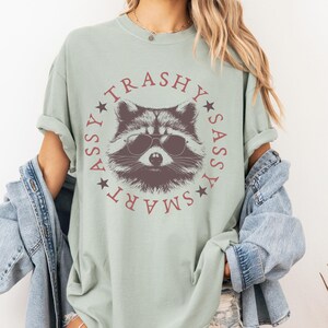 Puede incluir: Una camiseta verde claro con un mapache con gafas de sol en el centro. El mapache est&aacute; rodeado de un c&iacute;rculo con las palabras "SMART", "SASSY", "TRASHY" y "ASSY" escritas alrededor.