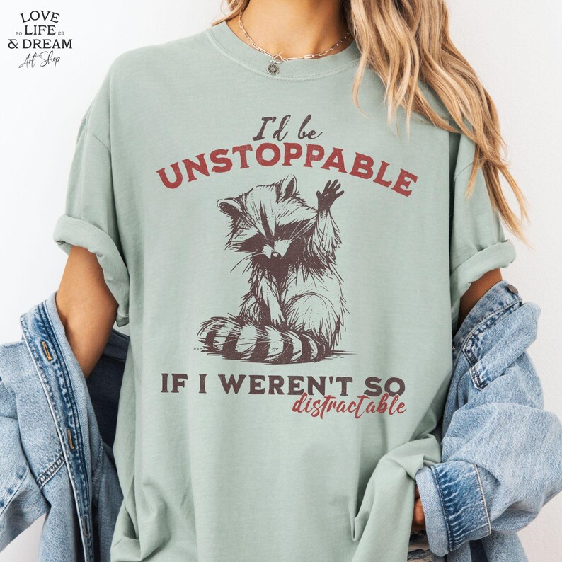 Może przedstawiać: Jasnozielona koszulka z napisem "I'd be UNSTOPPABLE IF I WEREN'T SO distractable" i ilustracją szopa pracza. Szop pracz jest brązowo-biały, z podniesioną łapą. Koszulka ma podwinięte rękawy.