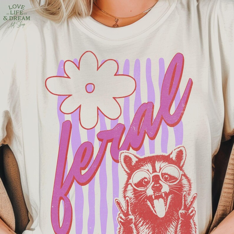 May include: Cream-colored t-shirt with a graphic design featuring a raccoon wearing sunglasses and giving the peace sign. The word "feral" is written in pink script, with a flower and purple stripes.
