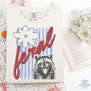 May include: Cream-colored t-shirt with a graphic of a raccoon wearing sunglasses and giving the peace sign, with the word "feral" in red script. The design includes a white flower and blue stripes. A delicate butterfly necklace is draped on the shirt.