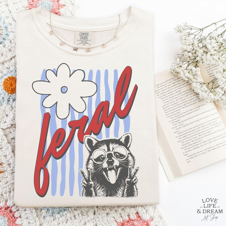 May include: Cream-colored t-shirt with a graphic of a raccoon wearing sunglasses and giving the peace sign, with the word "feral" in red script. The design includes a white flower and blue stripes. A delicate butterfly necklace is draped on the shirt.