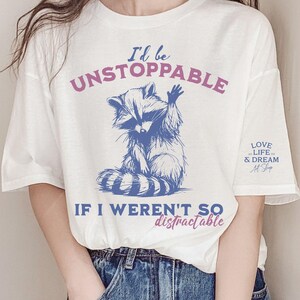 Może przedstawiać: Biała koszulka z niebieską ilustracją szopa pracza i napisem "I'd be UNSTOPPABLE IF I WEREN'T SO distractable." Na rękawie znajduje się r&oacute;wnież napis "LOVE LIFE & DREAM Art Shop".
