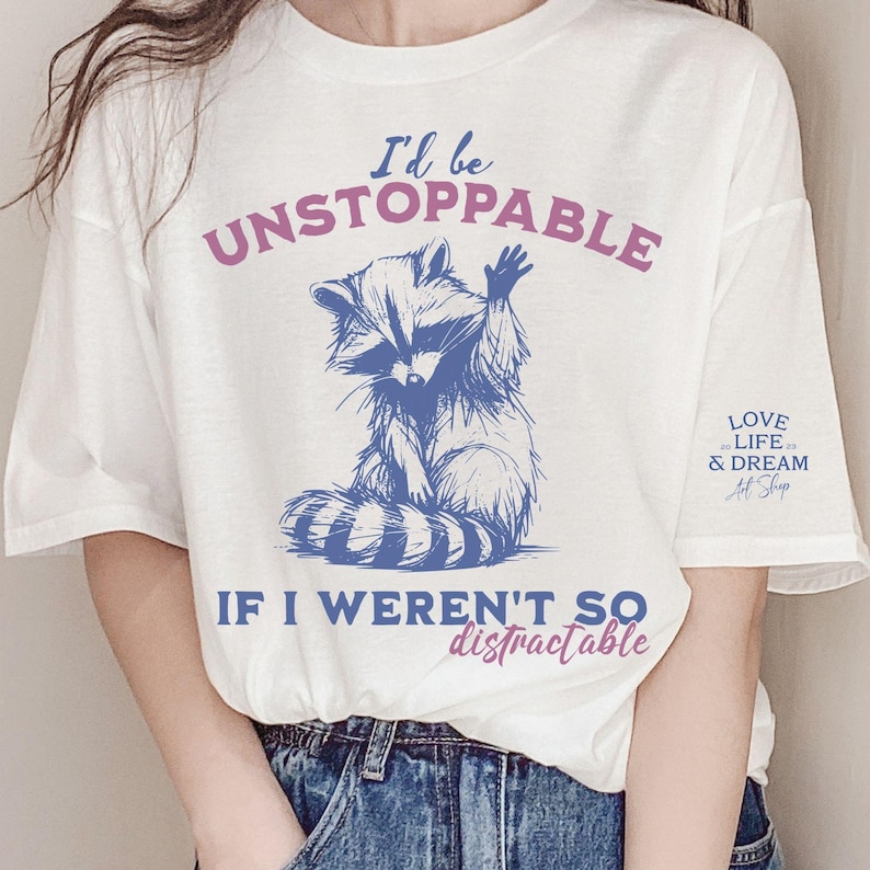 Może przedstawiać: Biała koszulka z niebieską ilustracją szopa pracza i napisem "I'd be UNSTOPPABLE IF I WEREN'T SO distractable." Na rękawie znajduje się r&oacute;wnież napis "LOVE LIFE & DREAM Art Shop".
