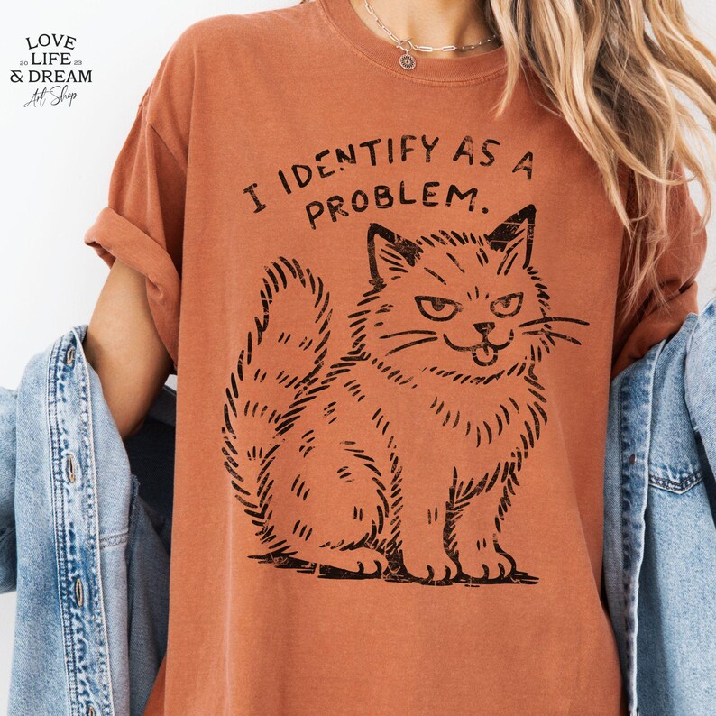 Puede incluir: Camiseta naranja quemado con el texto "I IDENTIFY AS A PROBLEM" y una ilustraci&oacute;n negra de un gato gru&ntilde;&oacute;n. La camiseta tiene mangas enrolladas y un corte relajado.