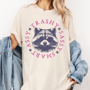Puede incluir: Una camiseta beige con un gr&aacute;fico de un mapache con gafas de sol. El mapache est&aacute; rodeado de un c&iacute;rculo con las palabras "SMART", "SASSY", "TRASHY" y "ASSY" en rosa.