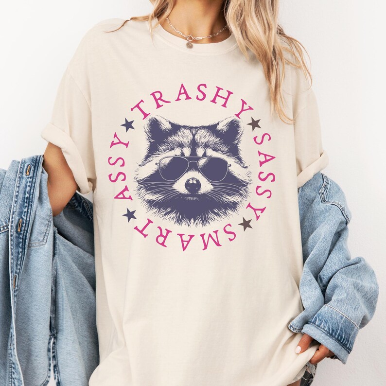 Puede incluir: Una camiseta beige con un gr&aacute;fico de un mapache con gafas de sol. El mapache est&aacute; rodeado de un c&iacute;rculo con las palabras "SMART", "SASSY", "TRASHY" y "ASSY" en rosa.