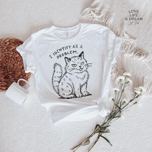 Puede incluir: Camiseta blanca con una ilustraci&oacute;n negra de un gato gru&ntilde;&oacute;n y el texto "I IDENTIFY AS A PROBLEM." El gato est&aacute; sentado con una expresi&oacute;n de suficiencia. La camiseta est&aacute; sobre una superficie blanca con una cesta tejida y una taza blanca.