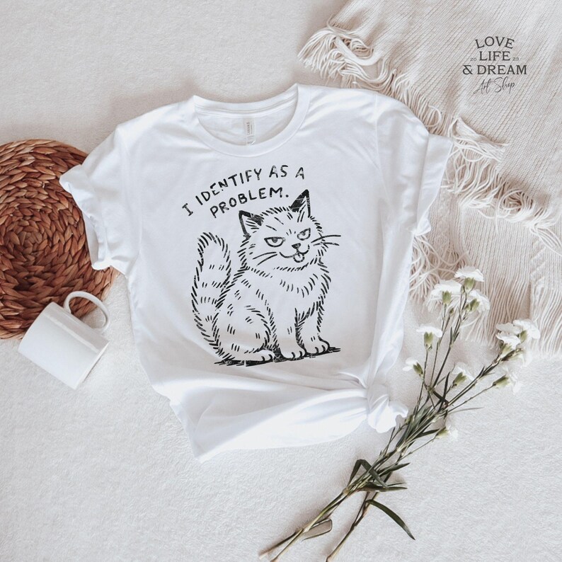 Puede incluir: Camiseta blanca con una ilustraci&oacute;n negra de un gato gru&ntilde;&oacute;n y el texto "I IDENTIFY AS A PROBLEM." El gato est&aacute; sentado con una expresi&oacute;n de suficiencia. La camiseta est&aacute; sobre una superficie blanca con una cesta tejida y una taza blanca.