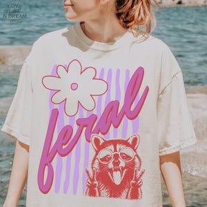 May include: Cream-colored t-shirt featuring a graphic of a raccoon wearing sunglasses and making a peace sign, with the word "feral" in pink script. A large flower and vertical purple stripes complete the design.