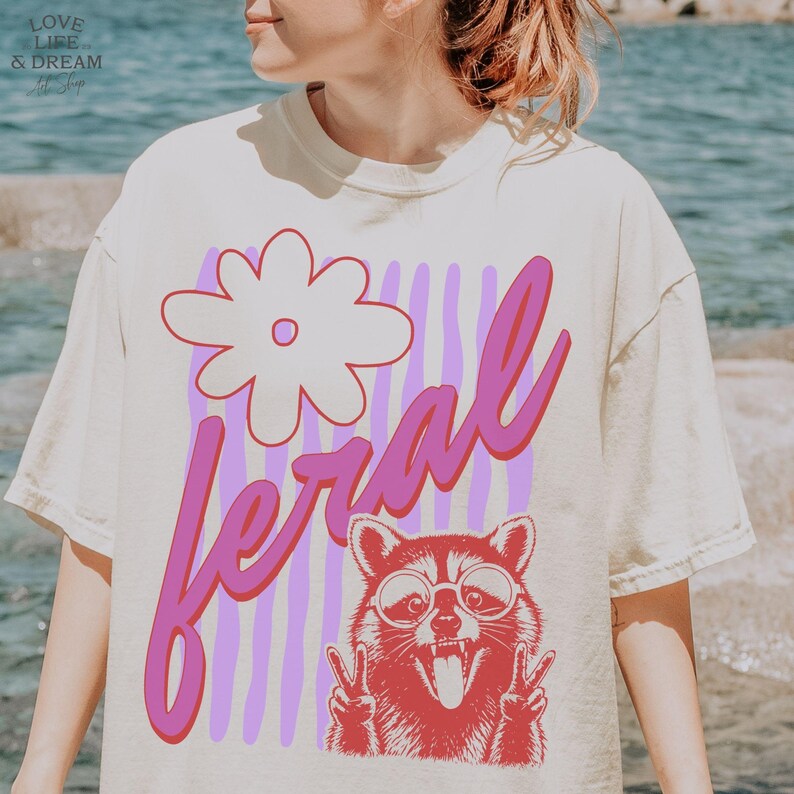 May include: Cream-colored t-shirt featuring a graphic of a raccoon wearing sunglasses and making a peace sign, with the word "feral" in pink script. A large flower and vertical purple stripes complete the design.