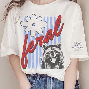 May include: White t-shirt with a graphic of a raccoon wearing glasses and making a peace sign. The word "feral" is in red script, with a white flower and blue stripes. The shirt also has the text "LOVE LIFE & DREAM Art Shop".