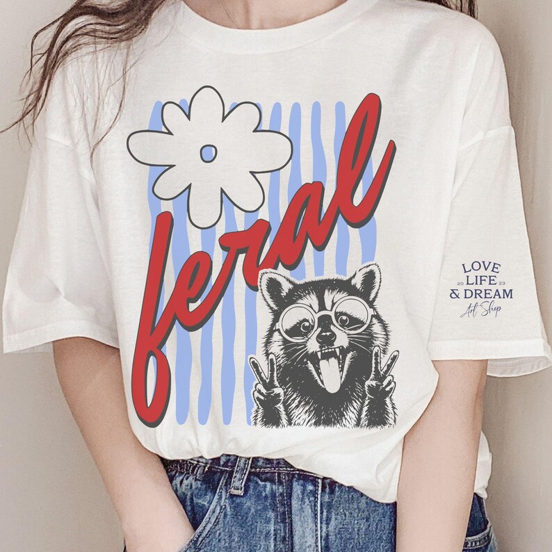May include: White t-shirt with a graphic of a raccoon wearing glasses and making a peace sign. The word "feral" is in red script, with a white flower and blue stripes. The shirt also has the text "LOVE LIFE & DREAM Art Shop".