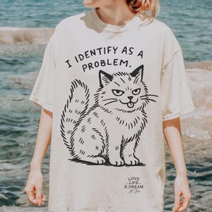 Puede incluir: Camiseta color crema con una ilustraci&oacute;n negra de un gato y el texto "I IDENTIFY AS A PROBLEM." El gato tiene una expresi&oacute;n gru&ntilde;ona. La camiseta tambi&eacute;n tiene el texto "LOVE LIFE & DREAM Art Shop."