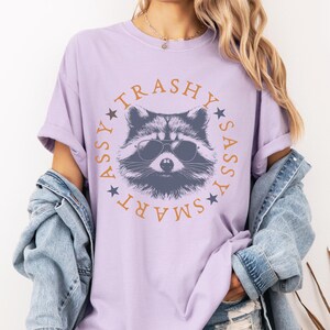 Puede incluir: Una camiseta lavanda con un gr&aacute;fico de un mapache con gafas de sol. El texto alrededor del mapache dice "SMART ASSY TRASHY SASSY".