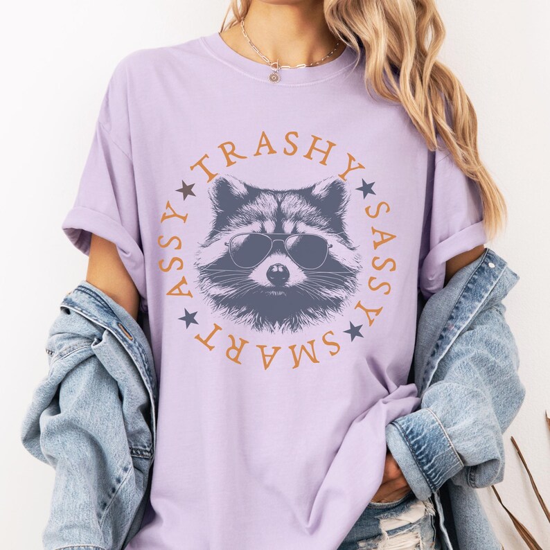 Puede incluir: Una camiseta lavanda con un gr&aacute;fico de un mapache con gafas de sol. El texto alrededor del mapache dice "SMART ASSY TRASHY SASSY".