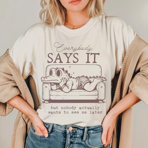 May include: A white t-shirt with a brown cartoon alligator lounging on a couch. The text "Everybody Says It" is above the alligator and the text "but nobody actually wants to see me later" is below the alligator.