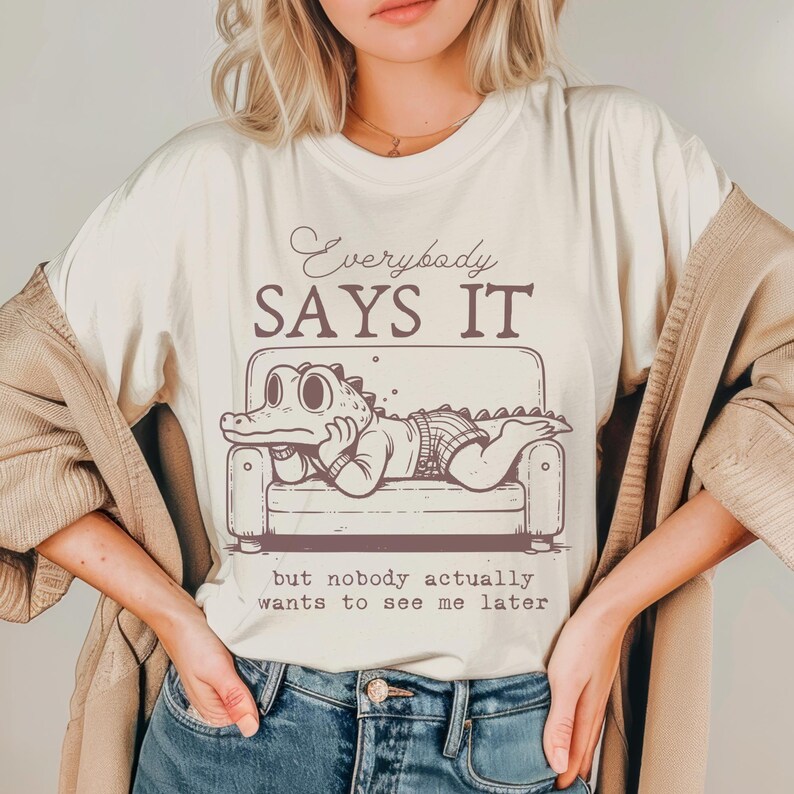 May include: A white t-shirt with a brown cartoon alligator lounging on a couch. The text "Everybody Says It" is above the alligator and the text "but nobody actually wants to see me later" is below the alligator.