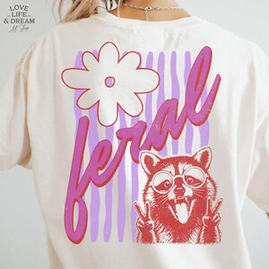 May include: Cream-colored t-shirt with a graphic design on the back. The design features a white flower, the word "feral" in pink, and a red raccoon wearing glasses and making a peace sign with its paws. The background has vertical purple stripes.