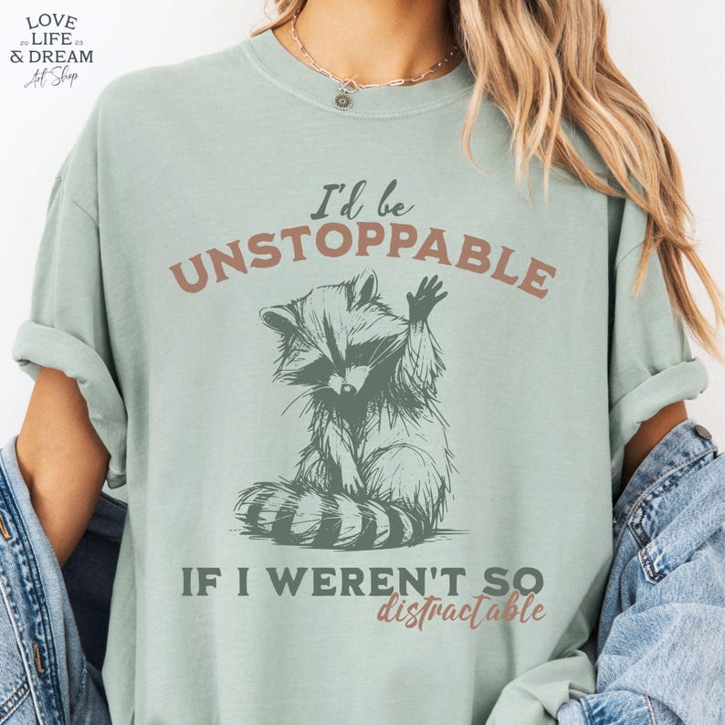 Może przedstawiać: Jasnozielona koszulka z grafiką szopa pracza i napisem "I'd be UNSTOPPABLE IF I WEREN'T SO distractable." Szop macha. Koszulka ma swobodny styl.