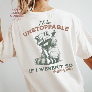 Może przedstawiać: Kremowa koszulka z grafiką szopa pracza i napisem "I'd be UNSTOPPABLE if I weren't so distractable." Szop pracz jest przedstawiony w szczeg&oacute;łowym stylu, z podniesionymi ramionami. Koszulka ma luźny kr&oacute;j.
