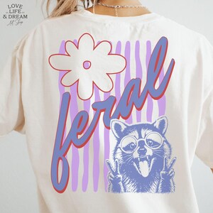 May include: Cream-colored t-shirt with a graphic design featuring a white flower, the word "feral" in blue and red, and a raccoon wearing glasses and making a peace sign.