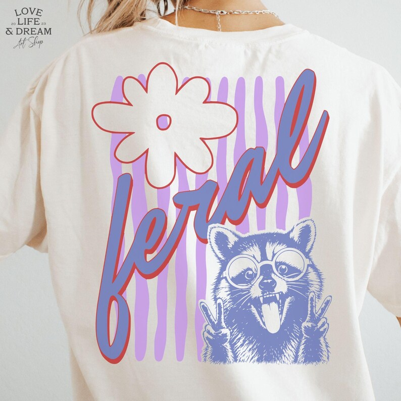 May include: Cream-colored t-shirt with a graphic design featuring a white flower, the word "feral" in blue and red, and a raccoon wearing glasses and making a peace sign.
