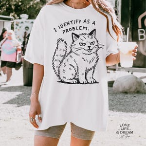 Puede incluir: Camiseta blanca con una ilustraci&oacute;n negra de un gato gru&ntilde;&oacute;n y el texto "I IDENTIFY AS A PROBLEM." El gato est&aacute; dibujado en estilo de dibujos animados con el ce&ntilde;o fruncido y la boca ligeramente abierta. La camiseta es holgada.