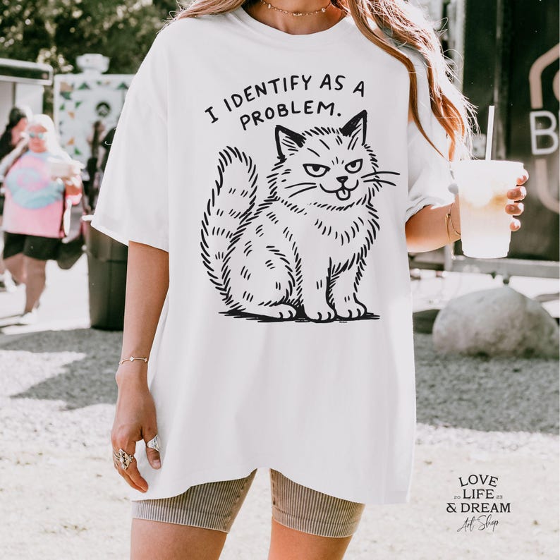 Puede incluir: Camiseta blanca con una ilustraci&oacute;n negra de un gato gru&ntilde;&oacute;n y el texto "I IDENTIFY AS A PROBLEM." El gato est&aacute; dibujado en estilo de dibujos animados con el ce&ntilde;o fruncido y la boca ligeramente abierta. La camiseta es holgada.