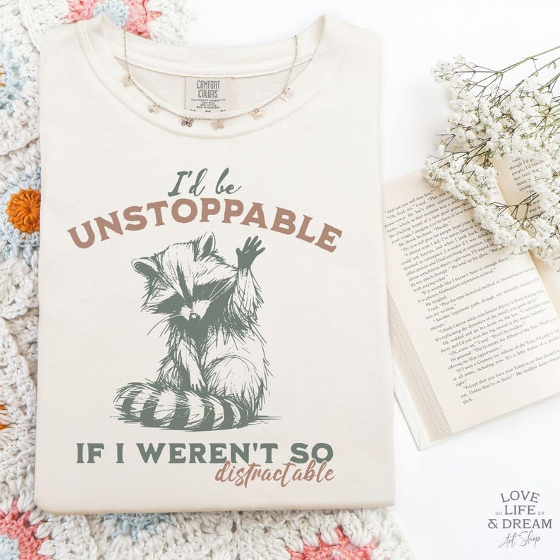 Może przedstawiać: Kremowa koszulka z napisem "I'd be UNSTOPPABLE IF I WEREN'T SO distractable" i ilustracją szopa pracza. Na koszulce leży delikatny srebrny naszyjnik z zawieszkami w kształcie motyli. Część kolekcji odzieży codziennej.