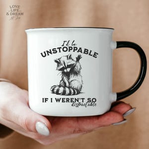 Może przedstawiać: Biały ceramiczny kubek z czarną obw&oacute;dką i uchwytem. Kubek przedstawia czarno-białą ilustrację szopa pracza z napisem "I'd be UNSTOPPABLE IF I WEREN'T SO distractable."