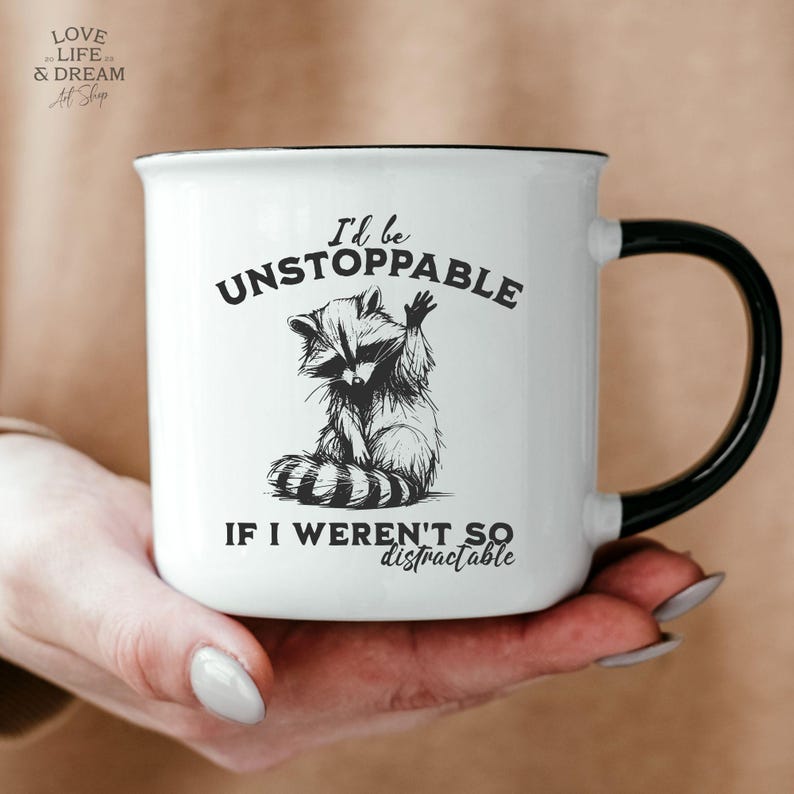 Może przedstawiać: Biały ceramiczny kubek z czarną obw&oacute;dką i uchwytem. Kubek przedstawia czarno-białą ilustrację szopa pracza z napisem "I'd be UNSTOPPABLE IF I WEREN'T SO distractable."