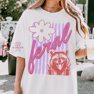 May include: White t-shirt with a graphic design featuring a raccoon wearing glasses and making peace signs. The word "feral" is written in pink script, with a flower and purple stripes. The text "LOVE LIFE & DREAM Art Shop" is on the left.
