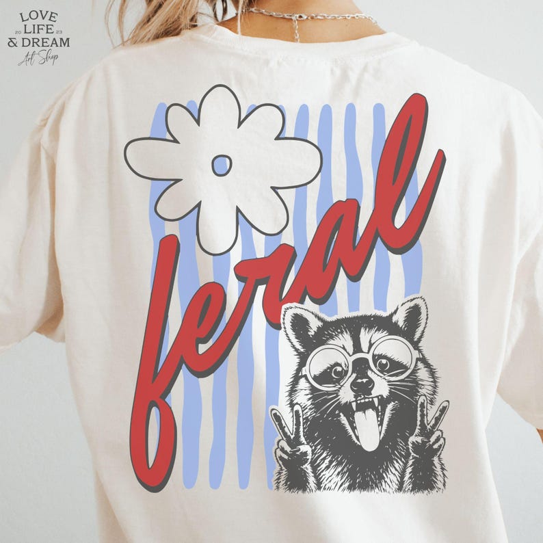 May include: Cream-colored t-shirt with a graphic design featuring a white flower, blue and white stripes, and the word "feral" in red script. A raccoon wearing glasses and making a peace sign is also featured.