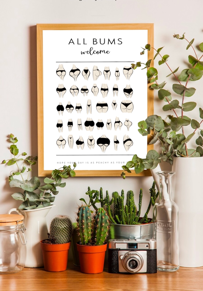 May include: A framed print with the text "ALL BUMS welcome" and a grid of 36 illustrations of different body shapes in black underwear. The text at the bottom reads "HOPE YOUR DAY IS AS PEACHY AS YOUR". The print is displayed on a shelf with potted plants and a camera.