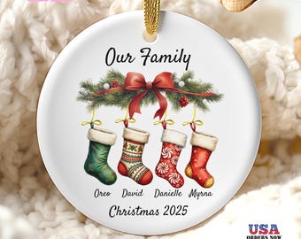 May include: A white ceramic Christmas ornament with a gold ribbon for hanging. The ornament features the text "Our Family" above four hanging stockings, each with a name. The bottom reads "Christmas 2025". A "Free Gift Box" label is in the upper left.