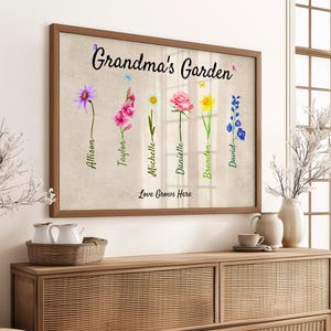 A Grandmas Garden sign featuring watercolor flowers and handwritten names is beautifully framed as a Grandmas Garden Framed Print. Below, the words Love Grows Here are elegantly inscribed. The artwork sits on a wooden sideboard, surrounded by decorative vases and lush plants.