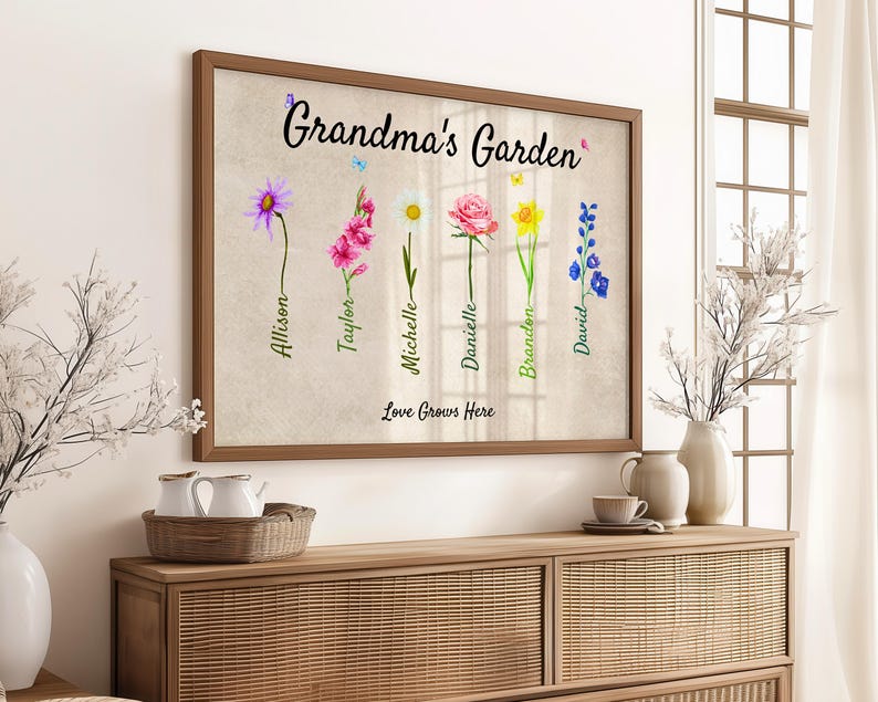 A Grandmas Garden sign featuring watercolor flowers and handwritten names is beautifully framed as a Grandmas Garden Framed Print. Below, the words Love Grows Here are elegantly inscribed. The artwork sits on a wooden sideboard, surrounded by decorative vases and lush plants.