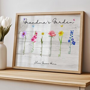 Op de afbeelding: Een wit houten bord met de tekst "Grandma's Garden" in zwarte cursieve letters. Het bord heeft een aquarel-illustratie van zes verschillende bloemen met namen onder elke bloem. De tekst "Love Grows Here" staat in zwarte cursieve letters onderaan het bord.