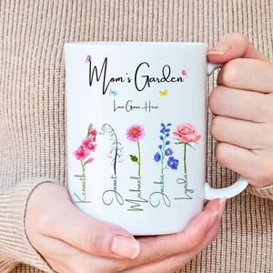 A person wearing a beige sweater holds a white 15 ounce mug adorned with colorful floral designs with kids names attached to the flower stems, bearing the text Mom's Garden followed by Love Grows Here in elegant cursive.