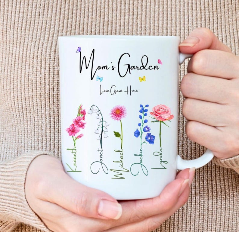A person wearing a beige sweater holds a white 15 ounce mug adorned with colorful floral designs with kids names attached to the flower stems, bearing the text Mom's Garden followed by Love Grows Here in elegant cursive.