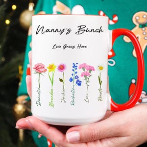 A person in a festive green sweater holds a white mug with a red handle. The mug reads “Nanny’s Bunch” and features illustrated flowers with handwritten names beneath each one.