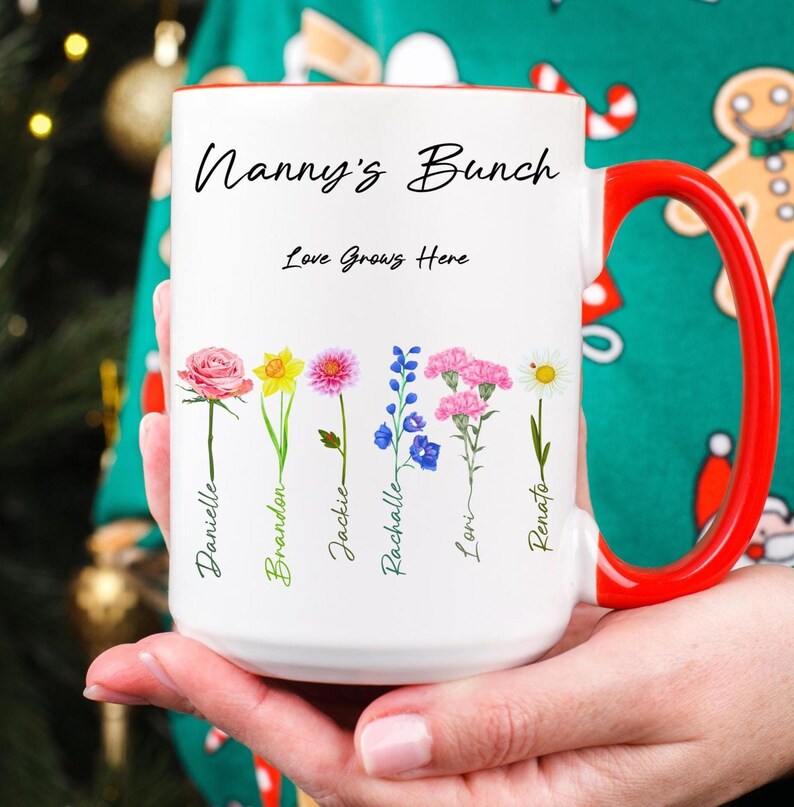 A person in a festive green sweater holds a white mug with a red handle. The mug reads “Nanny’s Bunch” and features illustrated flowers with handwritten names beneath each one.