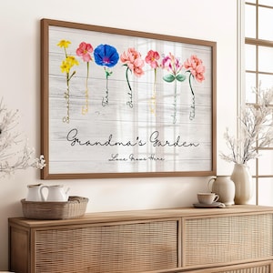 A Grandmas Garden sign featuring watercolor flowers and handwritten names is beautifully framed as a Grandmas Garden Framed Print. Below, the words Love Grows Here are elegantly inscribed. The artwork sits on a wooden sideboard, surrounded by decorative vases and lush plants.