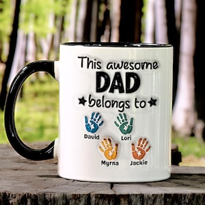 May include: White ceramic mug with a black handle and rim. The mug features the text "This awesome DAD belongs to" with handprint designs and names: David, Lori, Myrna, and Jackie. The mug is a personalized gift.